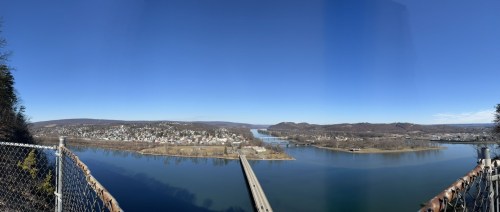 A panoramic view of a river with a bridge, surrounded by hills and a small town under a clear blue sky.