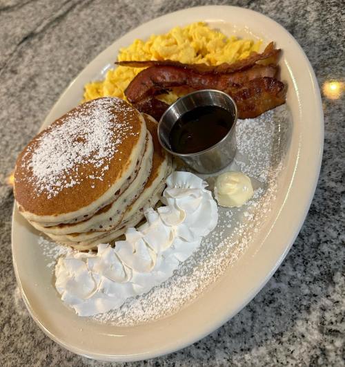 A plate of pancakes with whipped cream, syrup, scrambled eggs, and crispy bacon on a granite surface.