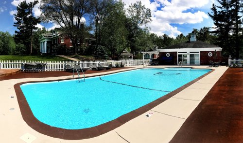 A bright outdoor pool surrounded by trees and a white fence, with a house and gazebo in the background under a blue sky.