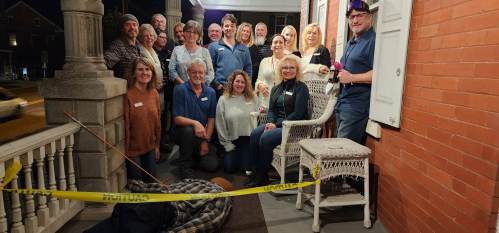 A group of people poses on a porch with caution tape and a person lying on the ground, suggesting a playful crime scene.