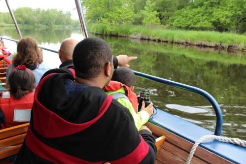 A man and child in life jackets point towards the water while on a boat, surrounded by greenery.