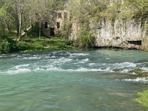 A serene river flows past a rocky bank and an old stone building, surrounded by lush greenery and trees.