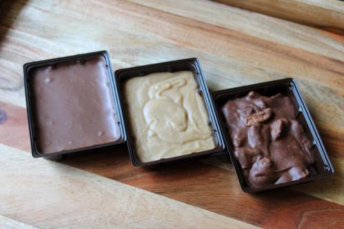 Three containers of different types of fudge: chocolate, caramel, and nutty chocolate, on a wooden surface.