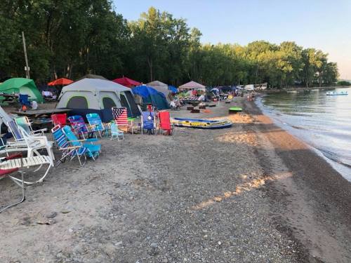A beach scene with tents, colorful chairs, and kayaks along the shore, surrounded by trees and calm water.