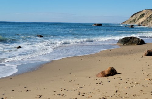 A serene beach scene with gentle waves, sandy shore, and rocky formations under a clear blue sky.