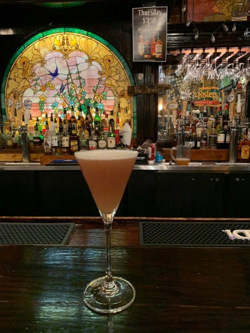 A cocktail in a martini glass sits on a bar, with a colorful stained glass backdrop and shelves of liquor bottles.