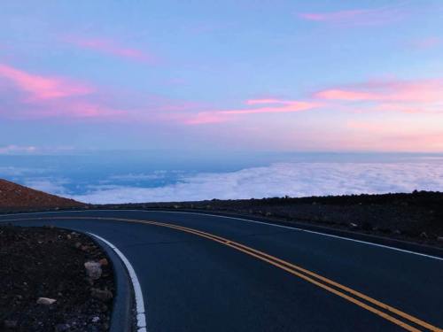 A winding road curves along a mountain, with a vibrant pink and blue sky above and clouds below.