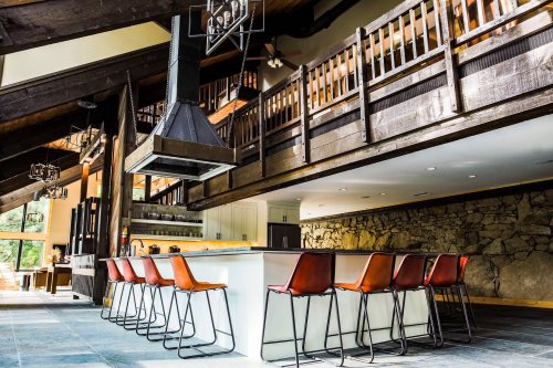 Modern kitchen with a large island and leather bar stools, featuring wooden beams and a stone wall in a spacious setting.