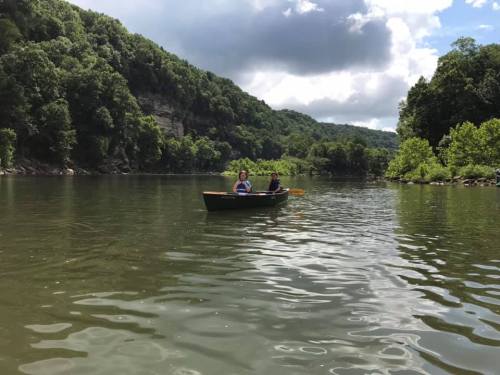 Two people in a small boat on a calm river, surrounded by lush green hills and a partly cloudy sky.
