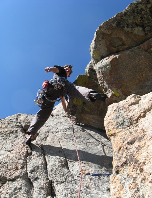 A climber ascends a rocky cliff, reaching for a handhold against a clear blue sky.