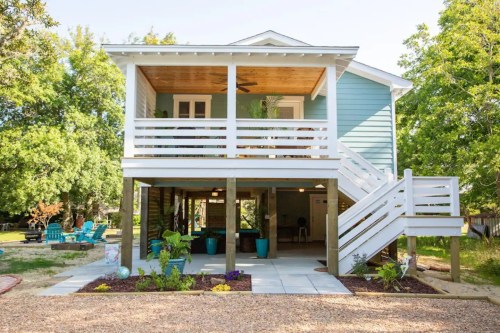 A two-story blue house with a porch, white railings, and a landscaped yard featuring plants and outdoor seating.