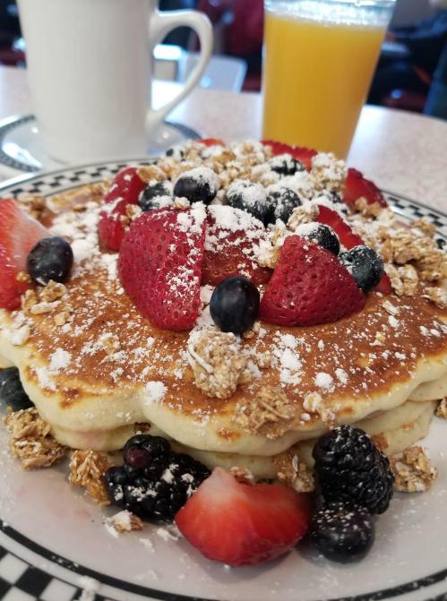 A stack of pancakes topped with strawberries, blueberries, blackberries, granola, and powdered sugar, with a drink in the background.