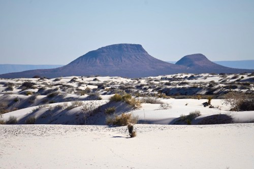 A vast desert landscape with white sand dunes and distant mountains under a clear blue sky.