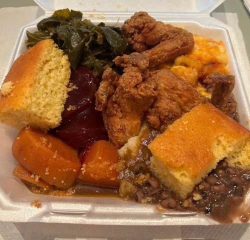 A takeout container filled with fried chicken, cornbread, collard greens, macaroni and cheese, and sweet potatoes.