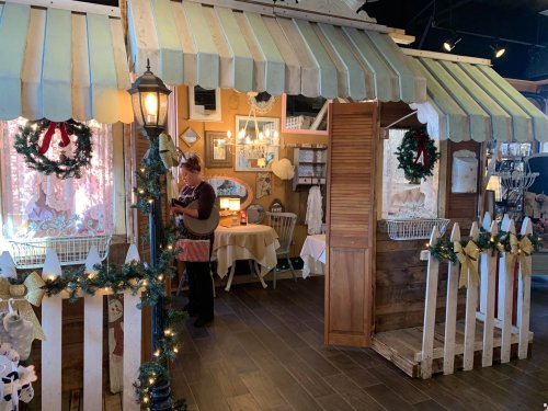 A cozy shop interior decorated for the holidays, featuring a woman arranging items and festive decor like wreaths and lights.