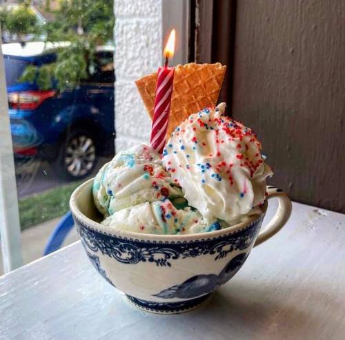 A bowl of ice cream topped with whipped cream, sprinkles, a candle, and a waffle cone, sitting by a window.