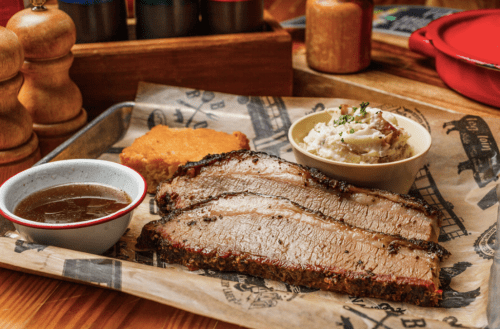 A plate featuring sliced brisket, creamy mashed potatoes, cornbread, and a small bowl of sauce on a rustic table.