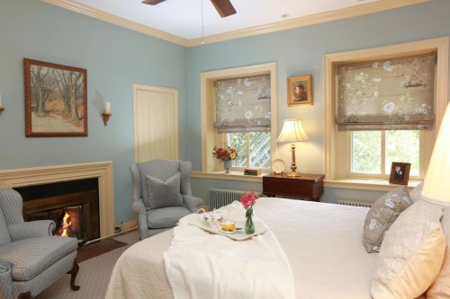 Cozy bedroom with a fireplace, blue walls, floral curtains, and a bed with a breakfast tray. Soft lighting and decor.