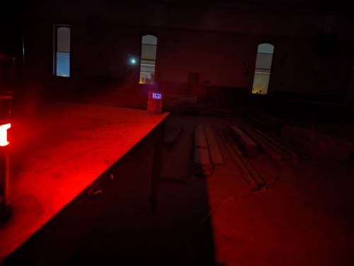 A dimly lit room with a red light, a small device on a table, and wooden planks scattered on the floor.