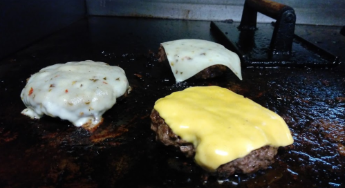 Three sizzling burgers on a grill, two topped with melted cheese and one with a slice of white cheese.