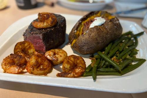 A plate featuring grilled shrimp, a steak, a baked potato with cheese, and green beans.