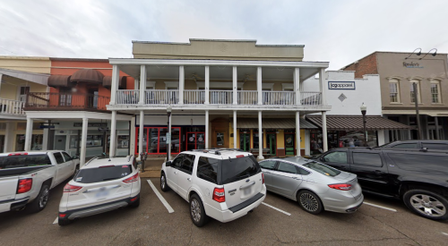 A two-story building with a balcony, surrounded by parked cars in a bustling street. Overcast sky above.