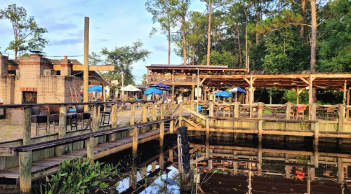 A rustic waterfront restaurant with outdoor seating, surrounded by trees and reflecting in the water.