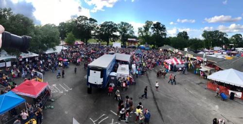 Aerial view of a crowded outdoor festival with colorful tents, food trucks, and people enjoying the event under a blue sky.