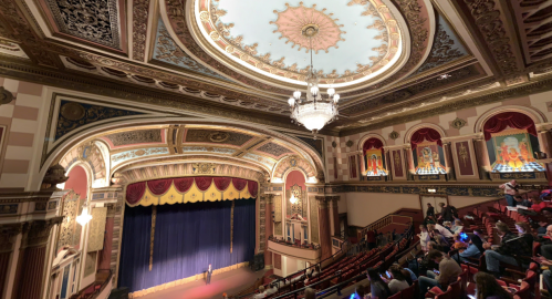 A grand theater interior with ornate ceiling, red curtains, and an empty stage, featuring elegant seating and decorative details.