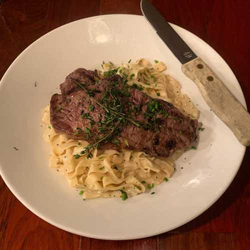 A plate of fettuccine topped with a grilled steak and garnished with herbs, alongside a knife.