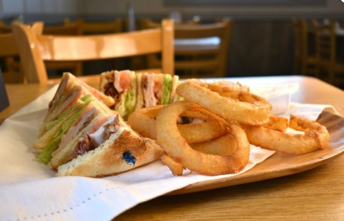 A plate with a club sandwich and crispy onion rings, set on a wooden table in a casual dining setting.