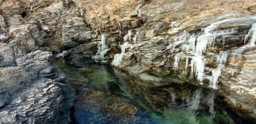 Rocky shoreline with icicles hanging from the cliffs, above a clear, greenish pool of water.