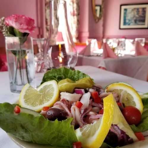A fresh salad with lettuce, lemon slices, olives, and diced vegetables, set on a table with a flower vase in the background.