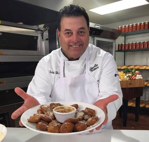A chef in a white coat proudly holds a large plate of golden-brown fried food with a dipping sauce in the center.