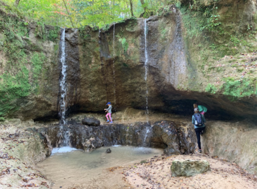 Two people explore a rocky area with a small waterfall and pool, surrounded by lush greenery.