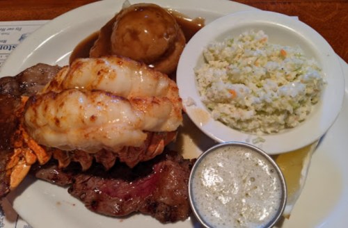 A plate featuring grilled lobster tail, steak, coleslaw, and a side of gravy with a biscuit.