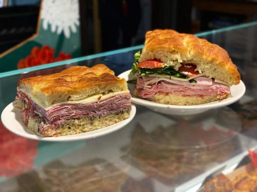 Two hearty sandwiches on plates, one sliced to reveal layers of meats, cheese, and fresh vegetables on a soft bread.