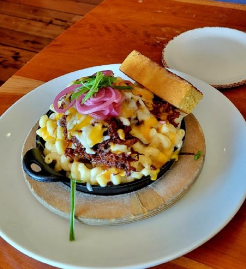A skillet of macaroni and cheese topped with pulled meat, cheese, pickled onions, and a slice of cornbread.