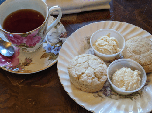 A floral teacup filled with tea beside a plate of three scones and two small cups of cream.