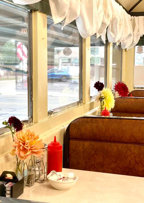 A cozy diner interior with flower centerpieces, ketchup bottle, and a view of the street through large windows.