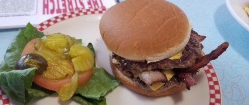 A burger with cheese and bacon on a bun, served with a side of lettuce, tomato, pickles, and olives.