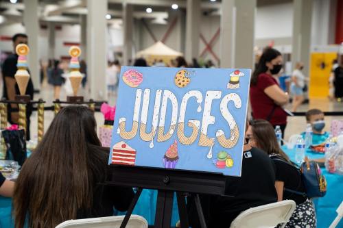 A colorful sign reading "JUDGES" displayed at a table in a busy event space, with people in the background.