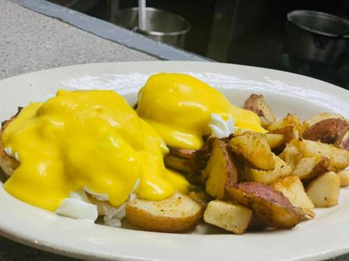 A plate of eggs Benedict topped with creamy yellow sauce, served with crispy potato wedges.