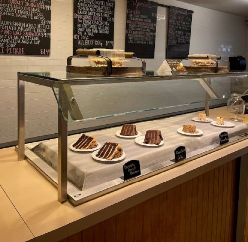 A display case with various cakes and desserts on white plates, with a chalkboard menu in the background.