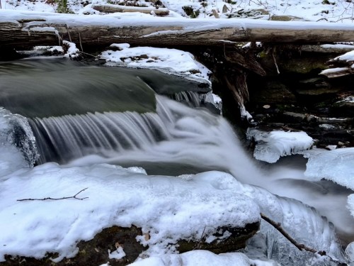 A serene winter scene featuring a flowing stream with ice and snow, framed by a fallen log and rocky terrain.