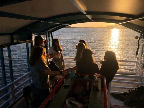 A group of people enjoying drinks on a boat at sunset, with the sun reflecting on the water.