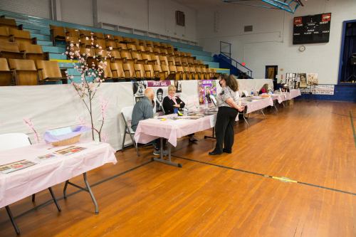 A gymnasium with tables set up for an event, featuring displays and attendees engaging in conversation.