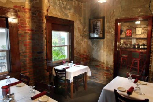 Cozy restaurant interior with exposed brick walls, wooden tables set for dining, and a bar visible in the background.