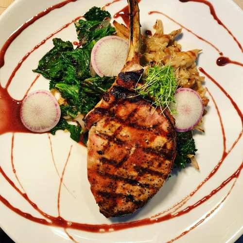 Grilled pork chop on a plate with sautéed greens, cabbage, radish slices, and a drizzle of sauce.
