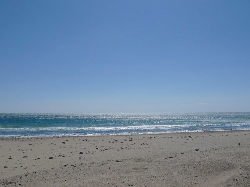 A serene beach scene with a clear blue sky, gentle waves, and a sandy shore dotted with small stones.